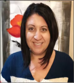 The image shows a woman with short hair smiling at the camera. She has a blue shirt with white sleeves, and there s an abstract background behind her.