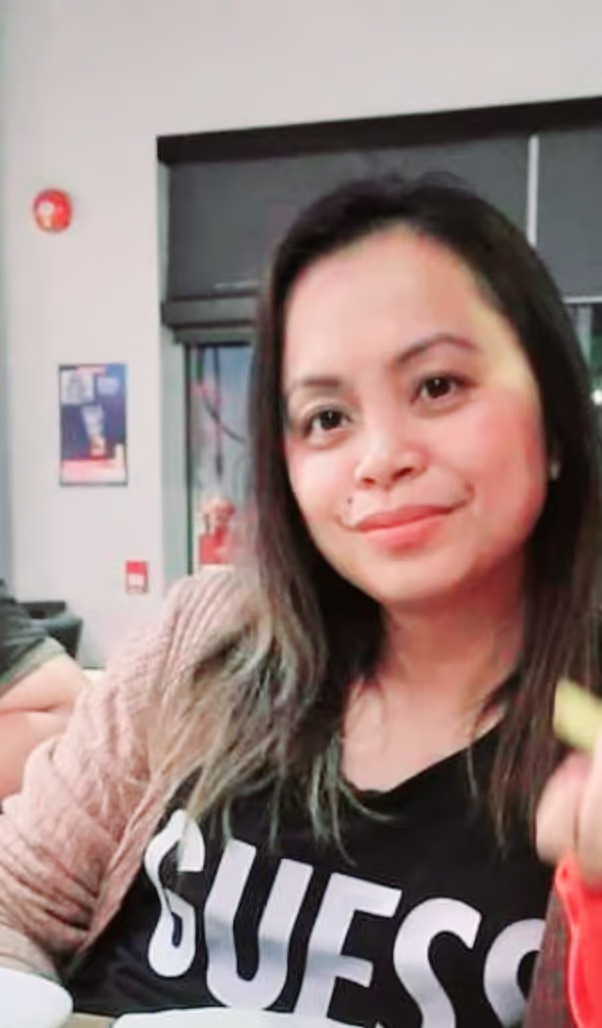 The image shows a woman with dark hair smiling at the camera, wearing a black shirt, seated indoors with a blurred background suggesting a casual dining setting.