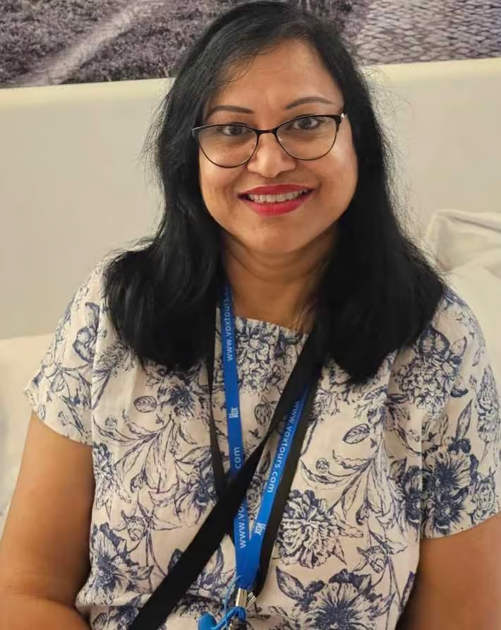 A woman with glasses, wearing a blue and white patterned shirt, sitting at a table with a lanyard around her neck, posing for a portrait.