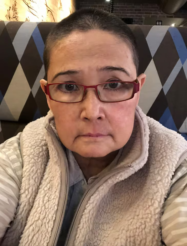 The image shows a person taking a selfie with a bald head wearing glasses, a red shirt, and a white jacket, sitting at a dining table with a blue and black striped backdrop.