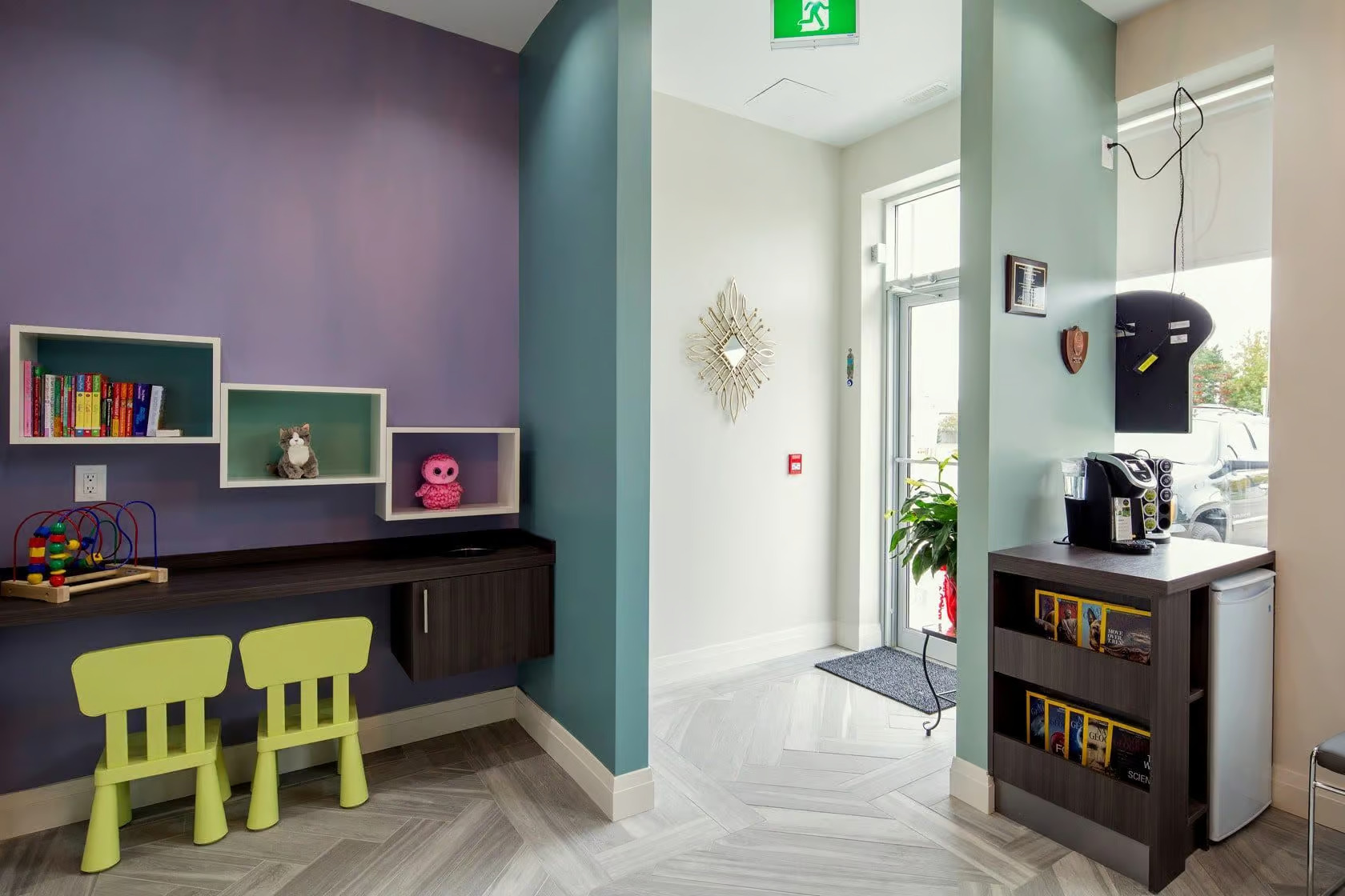 The image shows a room with purple walls and white flooring, featuring a play area with colorful furniture and decorations, a desk with shelves, a bookshelf, and a window with blinds partially drawn up.