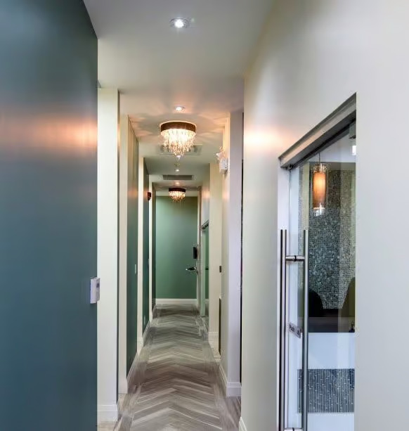 The image shows an interior hallway with a long corridor leading towards a door at the end, featuring modern decor elements such as chandeliers and contemporary wall finishes.