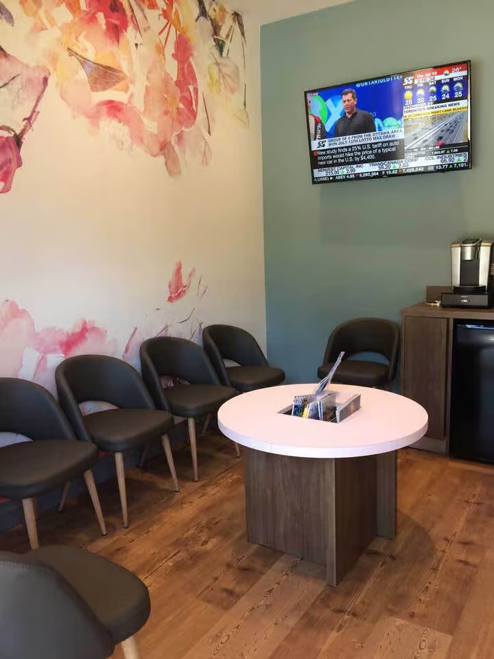 The image shows an interior space, likely a waiting area within a business establishment, featuring a seating arrangement with chairs around a small table, a television screen displaying news content, and a counter with a cash register.